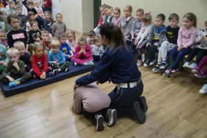 zdjęcie kolorowe zrobione w środku sali policjantka prezentuje wraz z uczeniem pozycję jaką należy przyjąć w przypadku kontaktu z niebezpiecznym psem reszta dzieci siedzi na ławkach