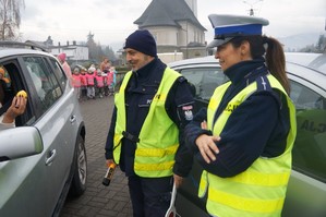 zdjęcie zrobione na zewnątrz widać fragment samochodu i rękę kierowcy która trzyma cytrynę obok stoi policjant z policjantką w tle dzieci