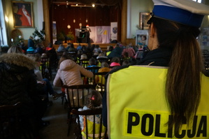 na zdjęciu policjantka stoi tyłem, w tle sala na krzesłach siedzą dzieci w tle scena