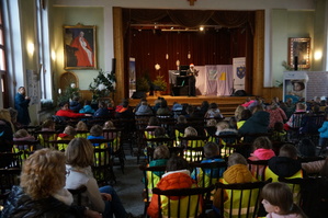 zdjęcie kolorowe zrobione w środku w sali na krzesłach tyłem siedzą dzieci przed nimi scena
