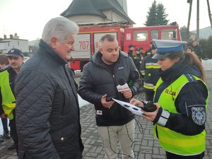 na zdjęciu kolorowym stoi policjantka redaktor radia oraz wójt gminy w czasie nagrania do radia w tle mieszkańcy