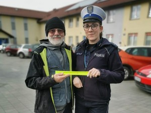 zdjęcie kolorowe zrobione na zewnątrz stoi policjantka obok niej mężczyzna który trzyma opaskę odblaskową za nimi parking z samochodami i budynki