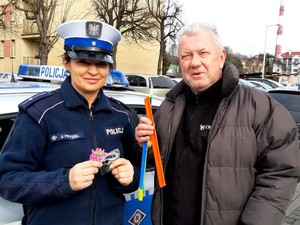 zdjęcie kolorowe zrobione na zewnątrz stoi policjantka obok niej mężczyzna oboje trzymają odblaskowe opaski w tle budynki i ulica