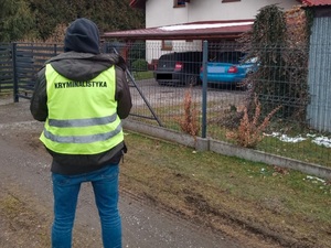 na zdjęciu kolorowym tyłem stoi technik kryminalistyki w tle budynek