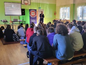 na zdjęciu kolorowym zrobionym w sali uczniowie siedzą tyłem przed nimi policjantka prowadzi prelekcje