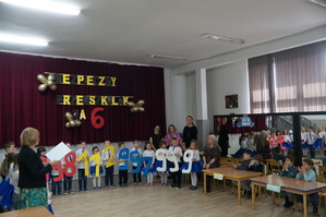 zdjęcie kolorowe zrobione w sali powitanie gości na środku stoją przedszkolaki trzymają wycięte z papieru numery alarmowe, uczestnicy siedzą przy stolikach