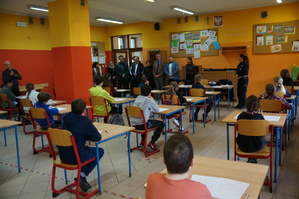 zdjęcie kolorowe zrobione w sali lekcyjnej za stolikami siedzą uczniowie przed nimi stoją organizatorzy i zaproszeni goście rozpoczęcie turnieju