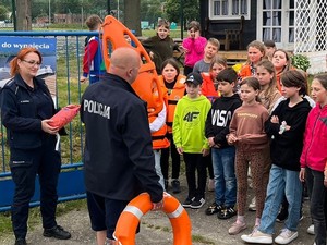 zdjęcie kolorowe policjant stoi tyłem i prowadzi prelekcje obok stoi policjantka przed nimi dzieci
