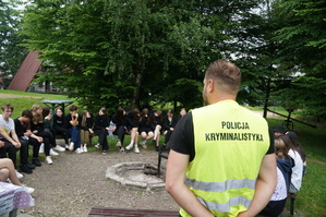 zdjęcie kolorowe tyłem stoi policjant ma założoną kamizelkę z napisem technik kryminalistyki wokoło siedzą uczniowie