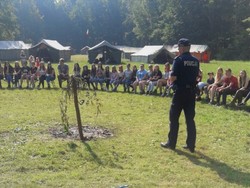 grafika poglądowa zdjęcie policjanta w czasie prelekcji w plenerze na ławkach siedzą harcerze