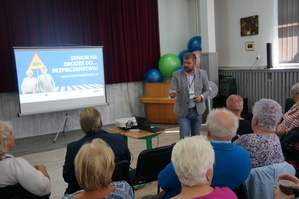 zdjęcie kolorowe zrobione w sali seniorzy siedzą tyłem przed nimi prowadzący