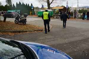 na pierwszym planie radiowóz w tle policjant stojący obok przejscia dla pieszych w tle cmentarz