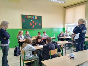 zdjęcie kolorowe zrobione w klasie uczniowie siedzą w ławkach i pracują w grupach obok stoi policjantka oraz nauczyciela
