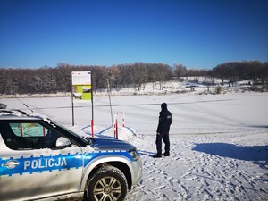 zdjęcie kolorowe na pierwszym planie radiowóz dalej policjant na tle zamarzniętego jeziora w tle góry
