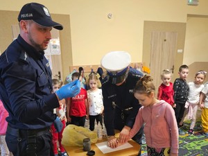 policjant tłumaczy jak zabezpiecza się linie papilarne obok policjantka i dziewczynka która odciska swoją dłoń na kartce