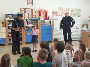 zdjęcie kolorowe policjantka prowadzi prelekcję obok niej stoi dziewczynka reszta dzieci siedzi