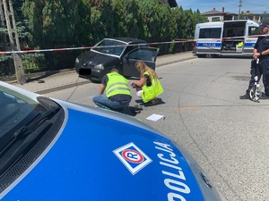 zdjęcie kolorowe na pierwszym planie radiowóz zw tle pracujący technik i policjant