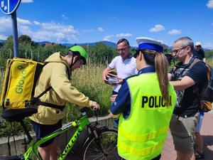 zdjęcie kolorowe przedstawiające policjantkę stojąca tyłem obok której stoi przedstawiciel Caritas którzy rozmawiają z rowerzystą