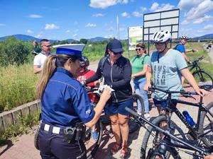 zdjęcie kolorowe przedstawiające policjantkę stojąca tyłem która przekazuje element odblaskowy młodemu rowerzyście.