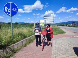 zdjęcie kolorowe przedstawiające policjantkę ruchu drogowego stojącą obok ścieżki rowerowej trzymająca plakat świetliści cykliści. obok niej stoi rowerzystka