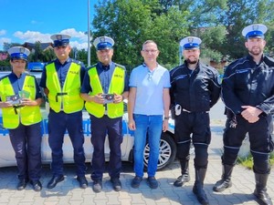 zdjęcie kolorowe przedstawiające stojących obok siebie trzech policjantów ruchu drogowego w kamizelkach odblaskowych, obok nich stoi przedstawiciel Caritas obok którego stoi dwoje policjantów w kombinezonach motocyklowych.