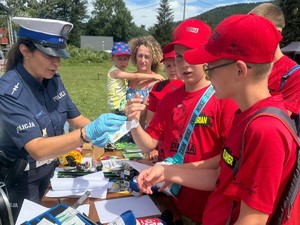 zdjęcie kolorowe przedstawiające policjantkę oraz dzieci stojące przy stoisku profilaktycznym. policjantka jest w rękawiczkach jednorazowych i przedstawia dzieciom jak się zbiera odciski palców.