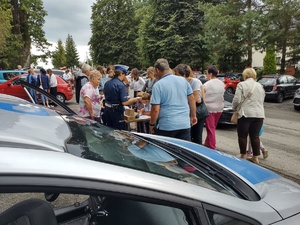 zdjęcie kolorowe na pierwszym planie fragment radiowozu w tle stoisko policyjna przy którym są osoby w tle zaparkowane samochody