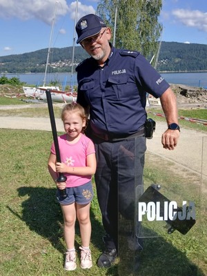 kolorowe zdjęcie policjanta obok niego stopi dziewczynka i trzyma elementy policyjnego wyposażenia