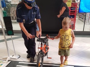 zdjęcie kolorowe przedstawiające policjantkę pokazującą dziecku jak bezpiecznie przeprowadzać rower na druga stronę jezdni