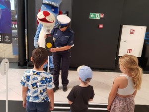 zdjęcie kolorowe przedstawiające policjantkę stojącą przy sygnalizatorze świetlnym, z przodu troje dzieci, z tyłu sznupek