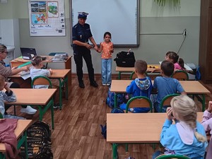 zdjęcie kolorowe zrobione w klasie uczniowie siedzą w ławkach policjantka z jedną uczennicą pokazuje jak się zachować w przypadku kontaktu z osobą obcą