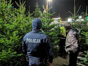 zdjęcie kolorowe policjant stoi tyłem obok niego mężczyzna który sprzedaje choinki, obok drzewka