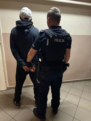 zdjęcie kolorowe, osoba zatrzymana w kajdanach, obok niego policjant