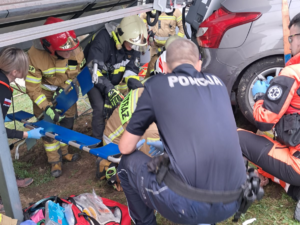 zdjęcie kolorowe, akcja ratunkowa, policjant kuca, dookoła strażacy