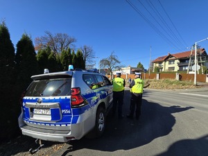 Na zdjęciu widoczny jest oznakowany radiowóz policyjny przy którym stoi dwóch policjantów ruchu drogowego
